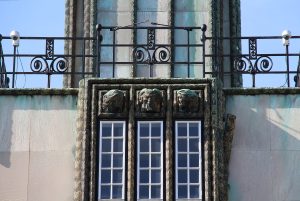 Decoration showing three carved heads above three slender vertical window openings. A balustrade with figural metlawork caps the wall.